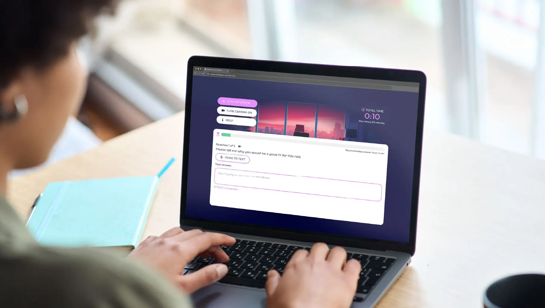 Person using a laptop to complete an AI interview simulator, with a virtual interview question displayed on screen, voice-to-text option enabled, and a countdown timer visible beside a notebook on the desk.